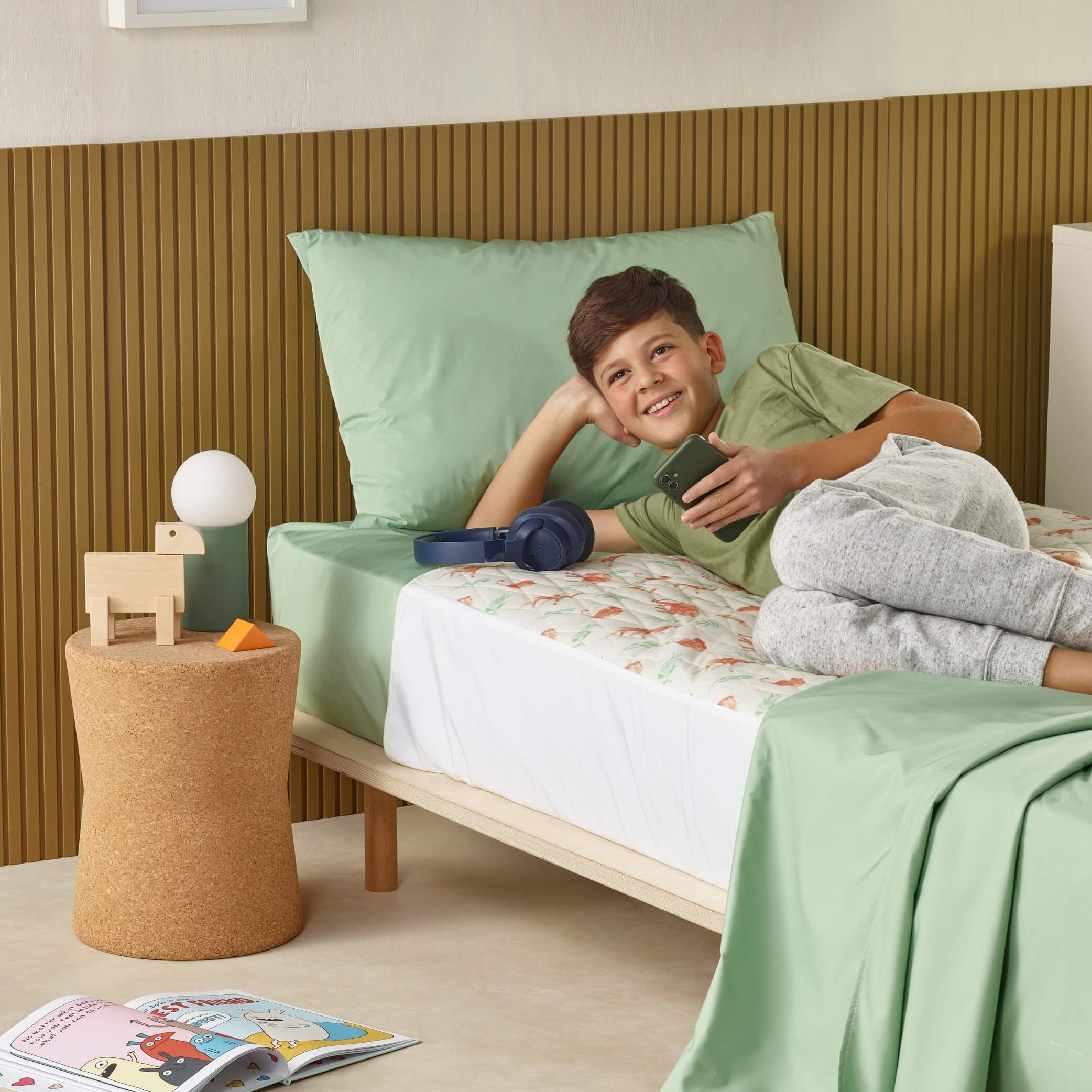 Boy relaxing with phone on bed with green waterproof bedding bed pad and sheets.